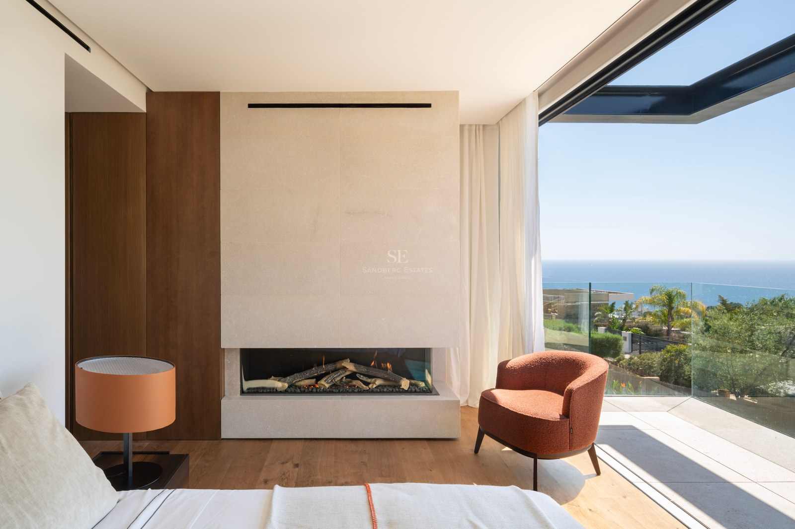 Chambre de maître moderne avec cheminée en pierre, fauteuil orange et baies vitrées ouvrant sur une terrasse vue mer.