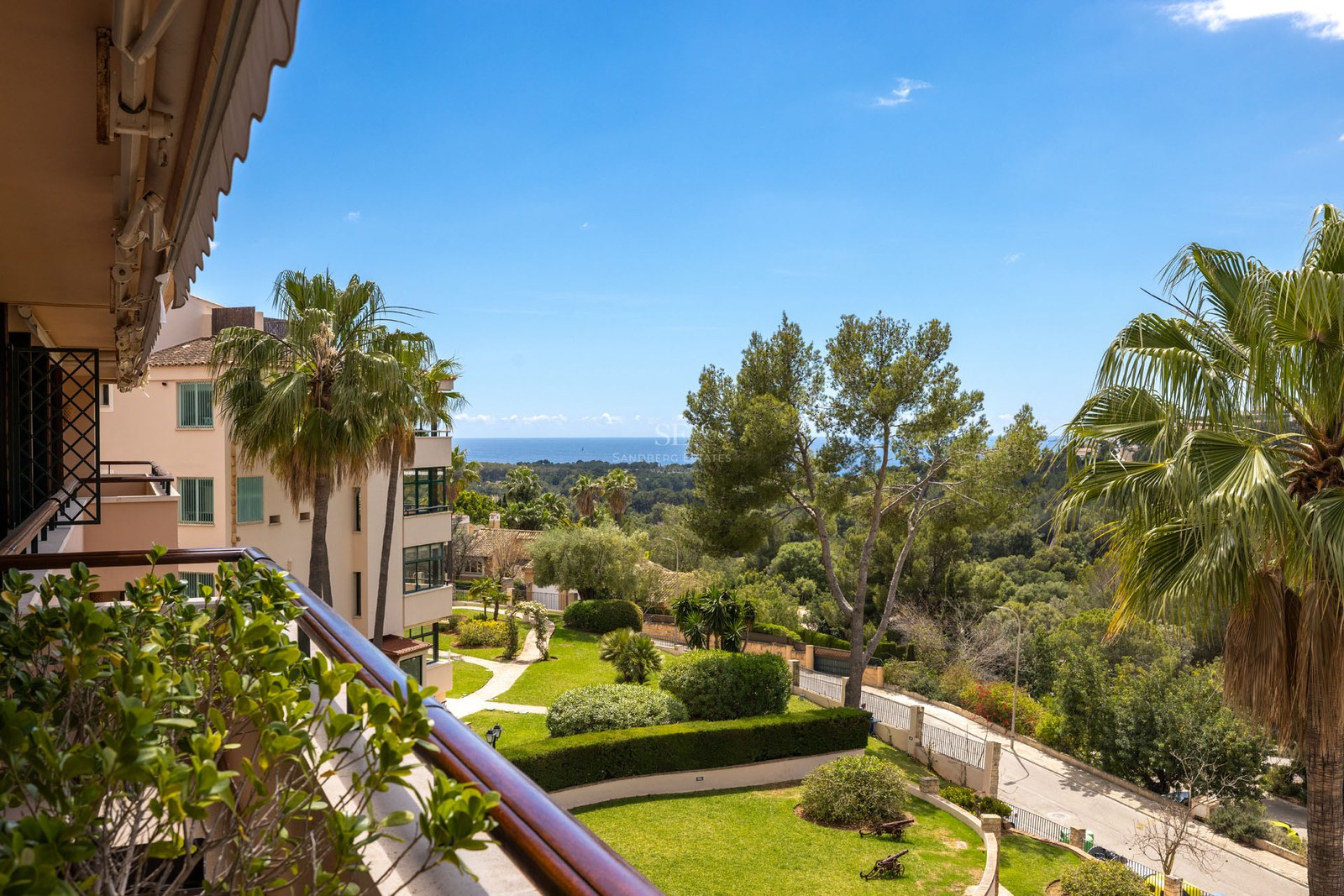 Vue d'un balcon sur un jardin méditerranéen avec des palmiers et la mer bleue au loin par temps clair.