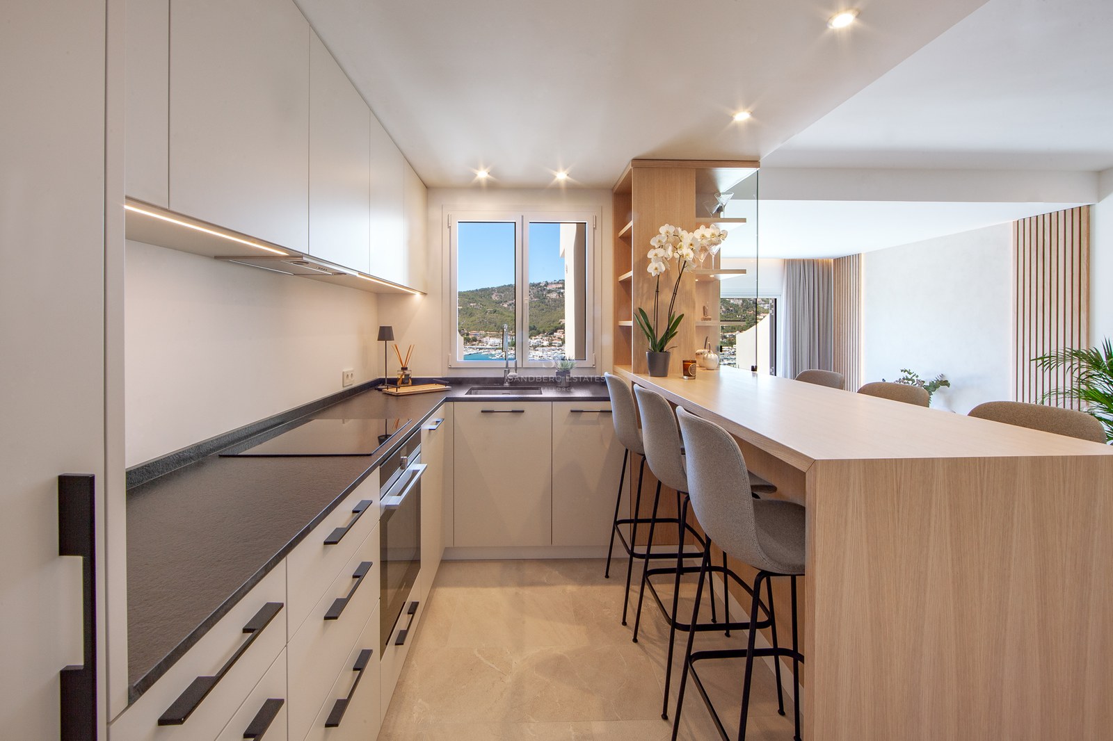 Modern kitchen with white cabinets, dark countertops, a wooden breakfast bar with three stools, and a window view of a harbor.