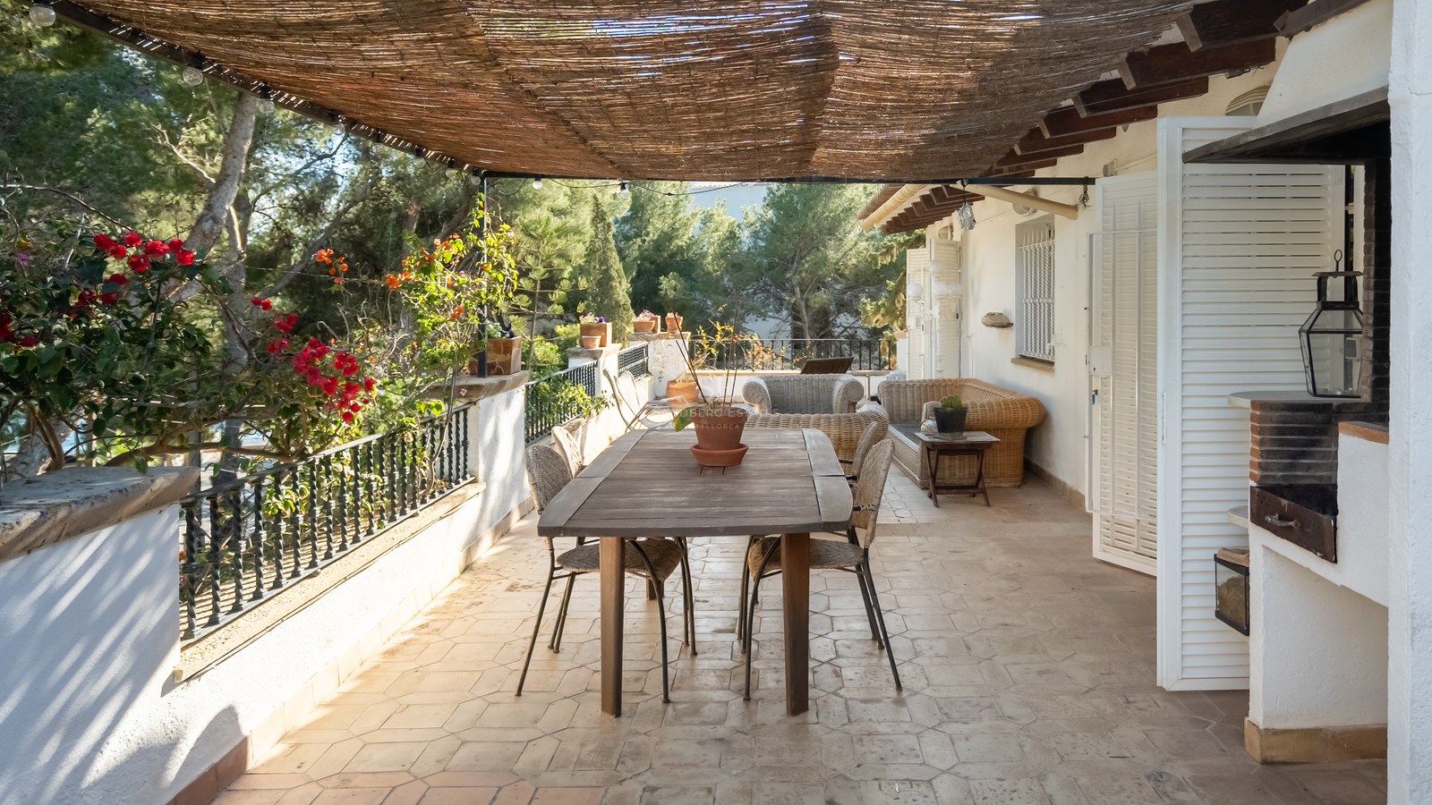Terraza sombreada con mesa de comedor de madera, muebles de mimbre y barbacoa integrada rodeada de vegetación.