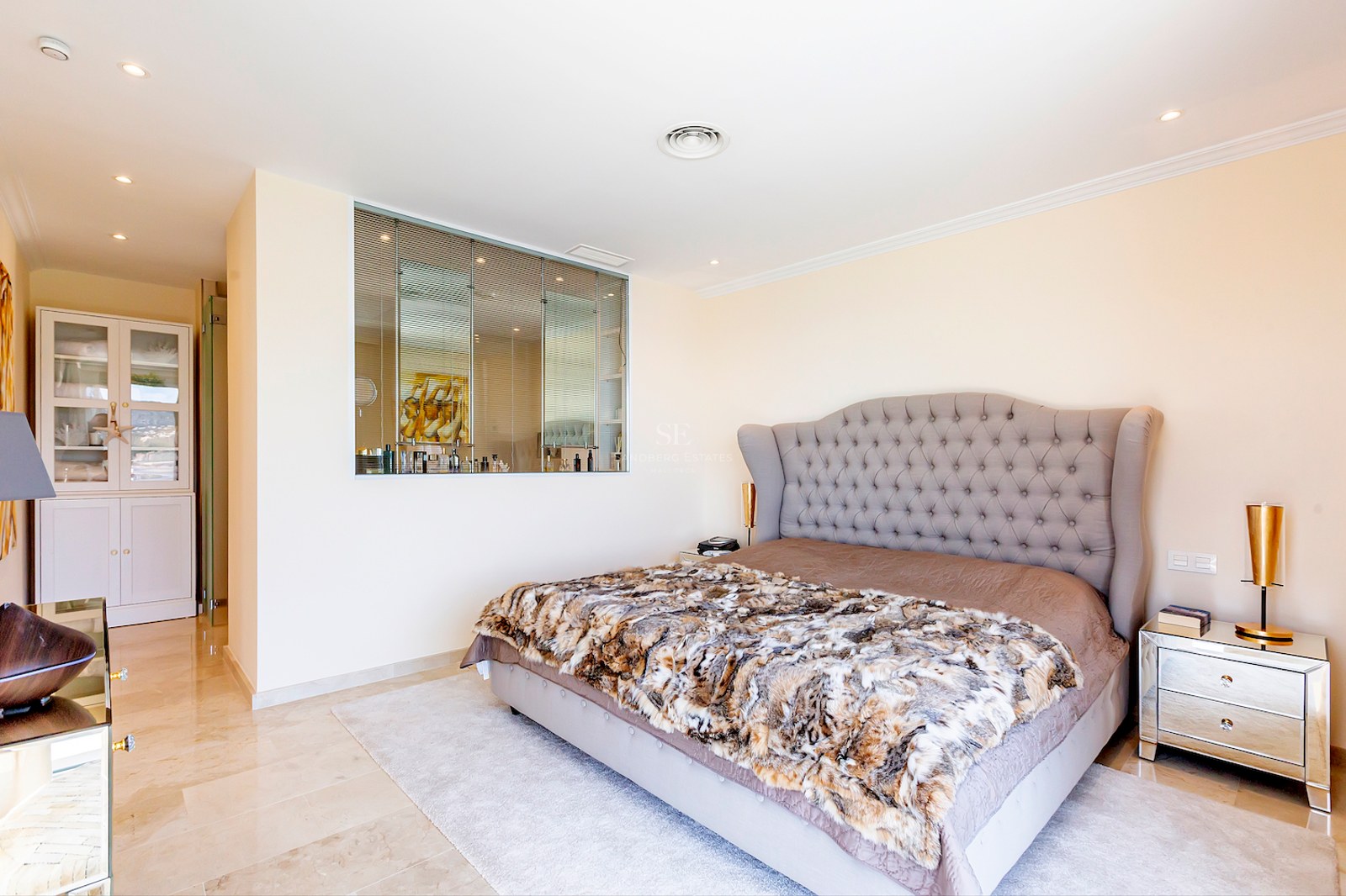 Spacious bedroom featuring a tufted grey headboard, marble floors, mirrored nightstands, and integrated storage.