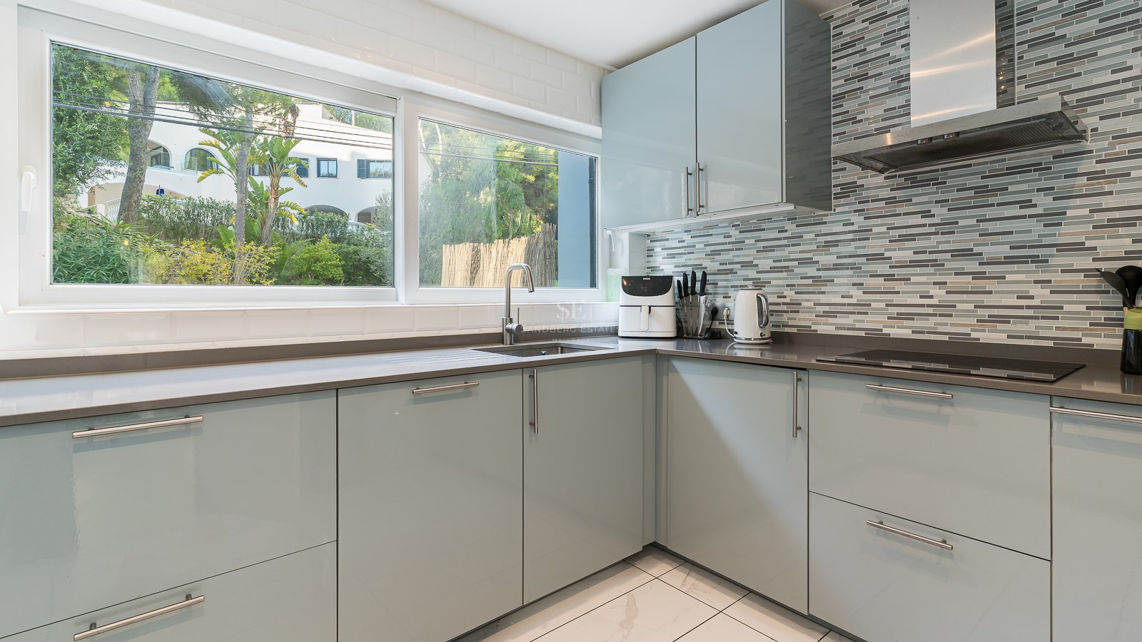 Modern light blue kitchen cabinets with grey countertops, a large window overlooking trees, and a mosaic tile wall.