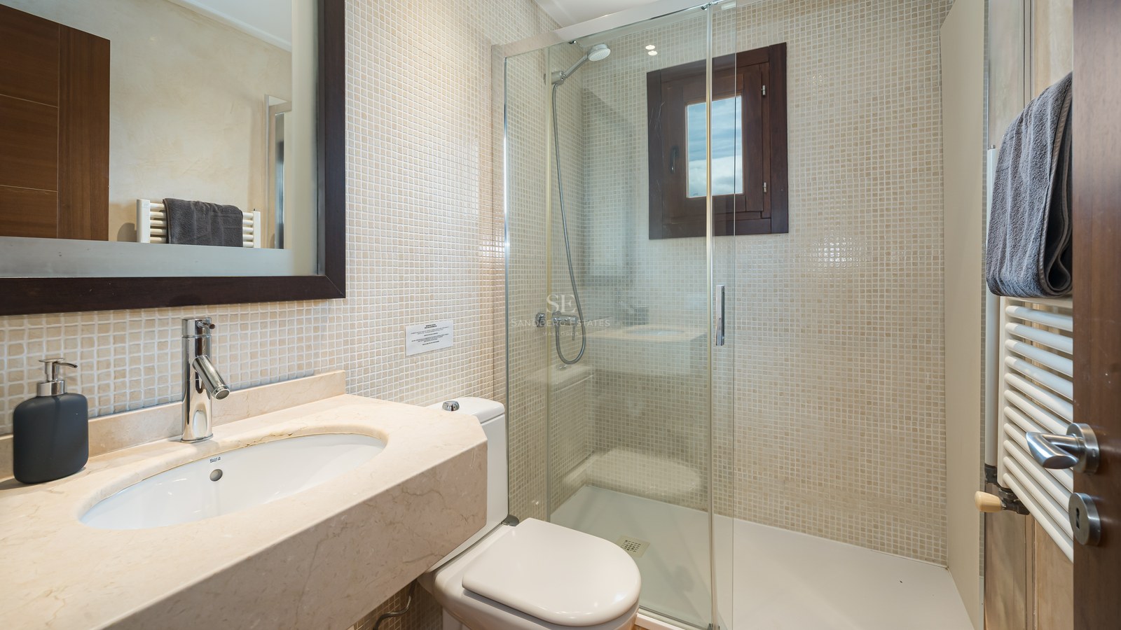 Badrum med glasdusch, beige mosaikväggar och tvättställ i marmor under en mörk spegelram.