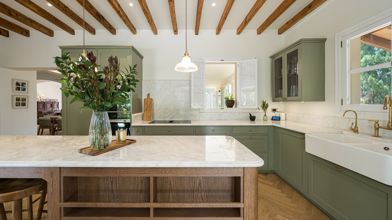 Modern sage green kitchen with marble countertops, wooden ceiling beams, and a large central island with flowers.
