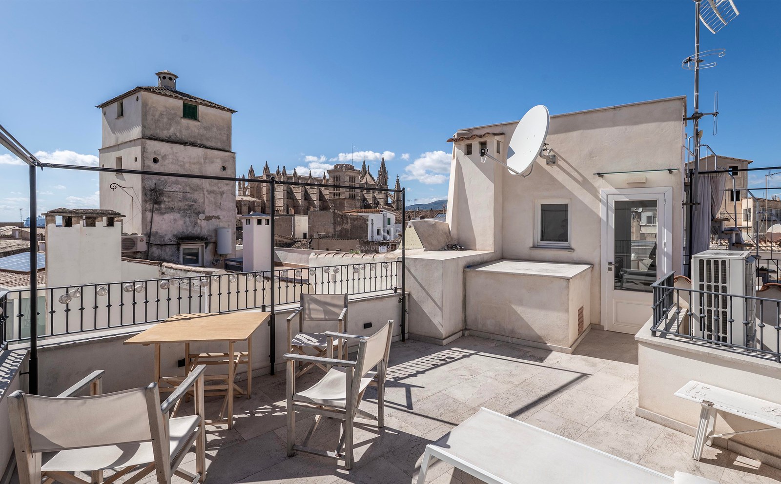 Spacious rooftop terrace with wooden furniture and a stunning view of Palma's historic cathedral under a clear blue sky.