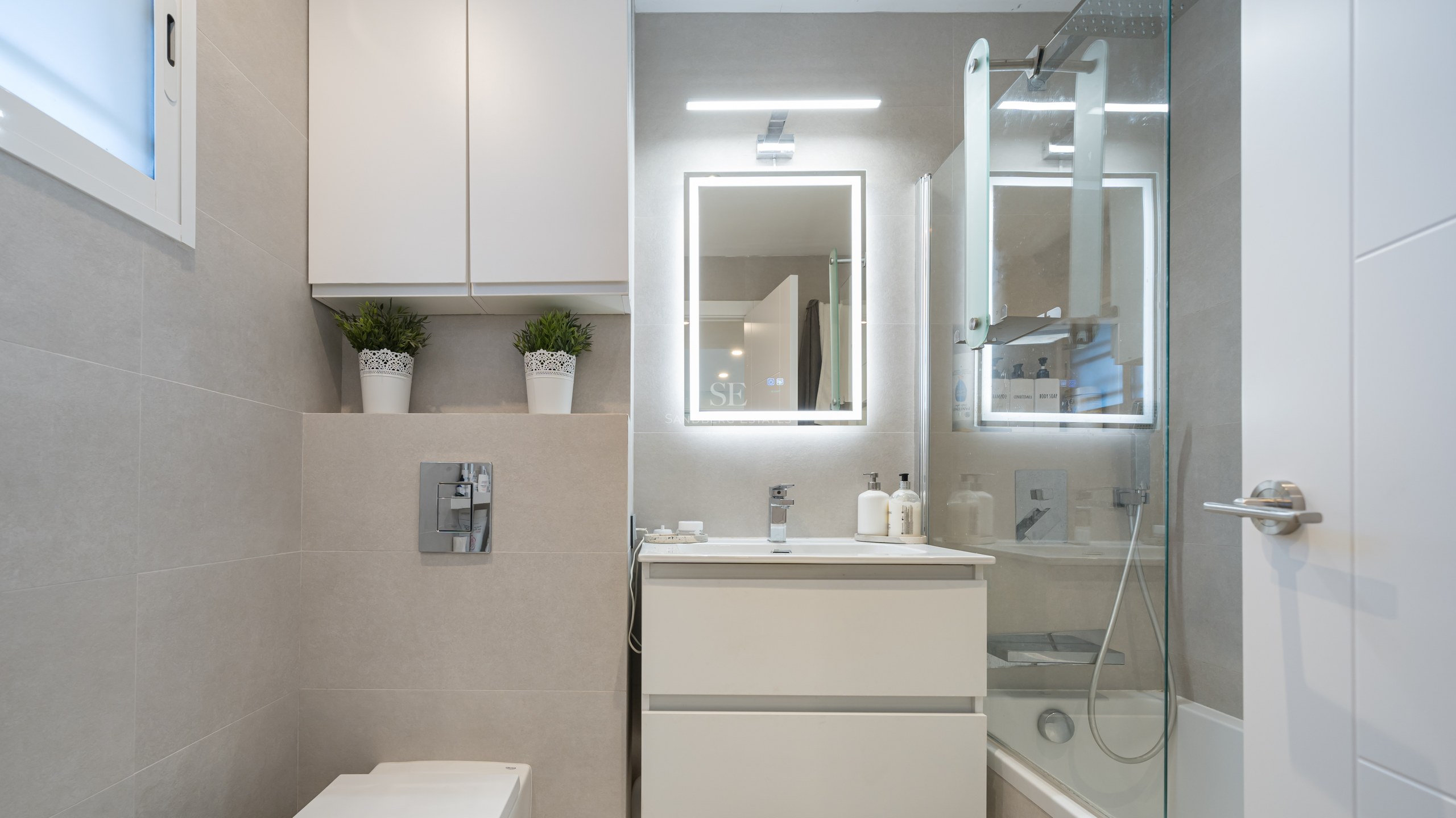Salle de bain moderne avec vanité blanche, miroir LED, paroi en verre et carrelage beige grand format.