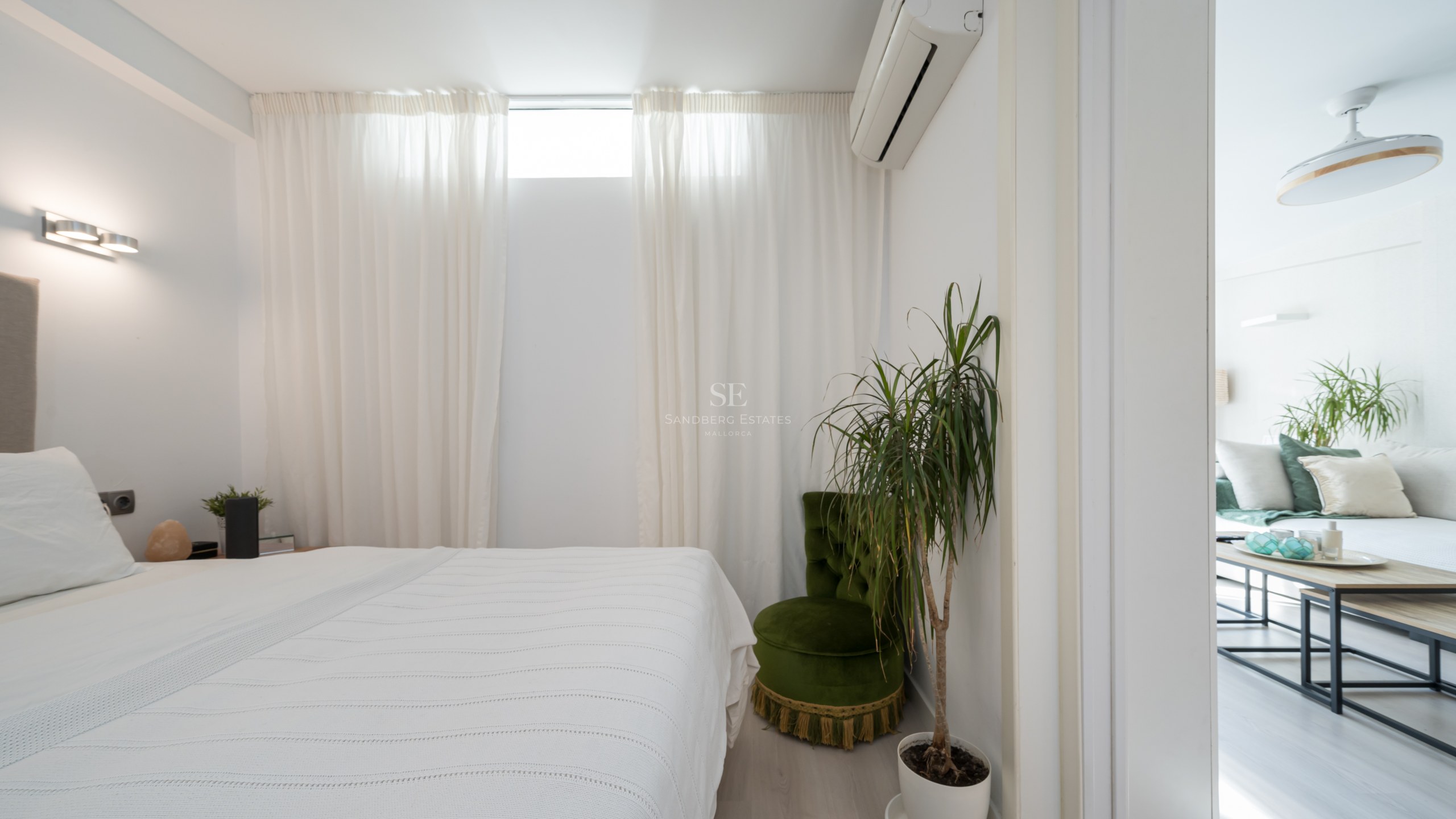 Chambre blanche épurée avec lit, fauteuil en velours vert, plante verte et vue sur le salon attenant.