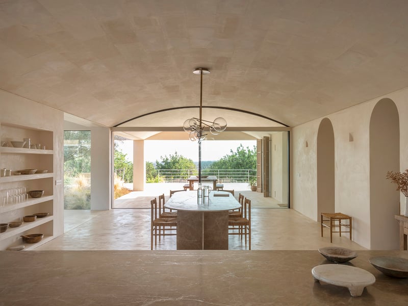 A minimalist dining room featuring a vaulted stone ceiling, oval marble table, and large glass doors to a sunlit terrace.