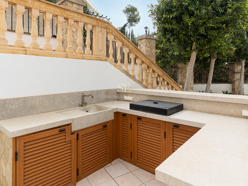 Cuisine extérieure avec plans de travail en pierre, meubles en bois et évier en laiton près d'une balustrade classique.