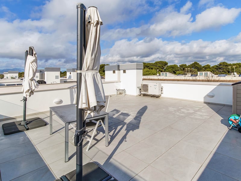 Grande terrasse sur le toit avec carrelage, table grise, chaises et parasols fermés sous un ciel nuageux.
