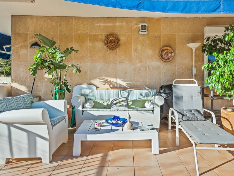 Terrasse ensoleillée avec mobilier de salon blanc, mur en pierre texturée et plantes vertes en pots.