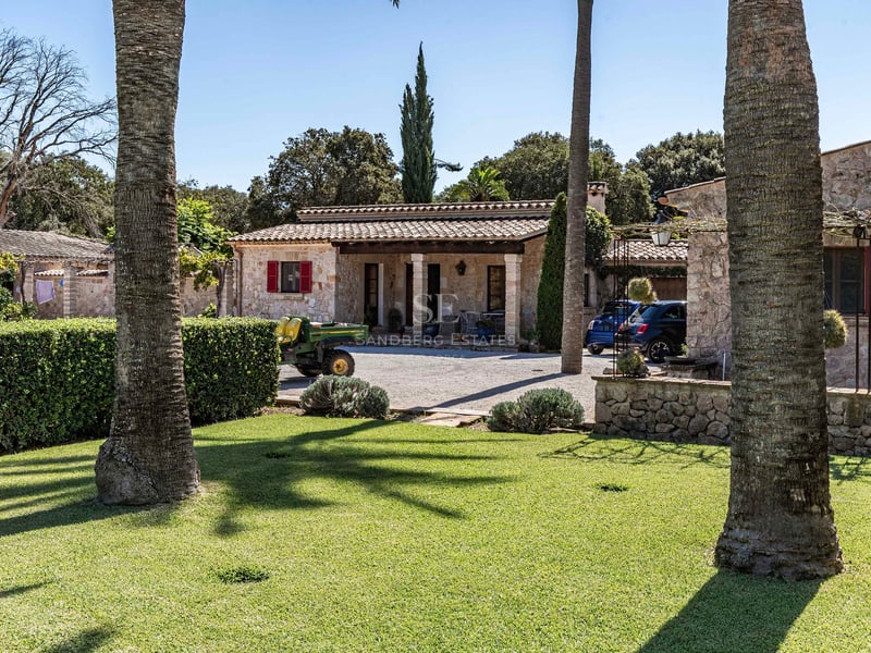 Villa rustique en pierre avec volets rouges et toit en tuiles, entourée d'une pelouse verdoyante et de palmiers.