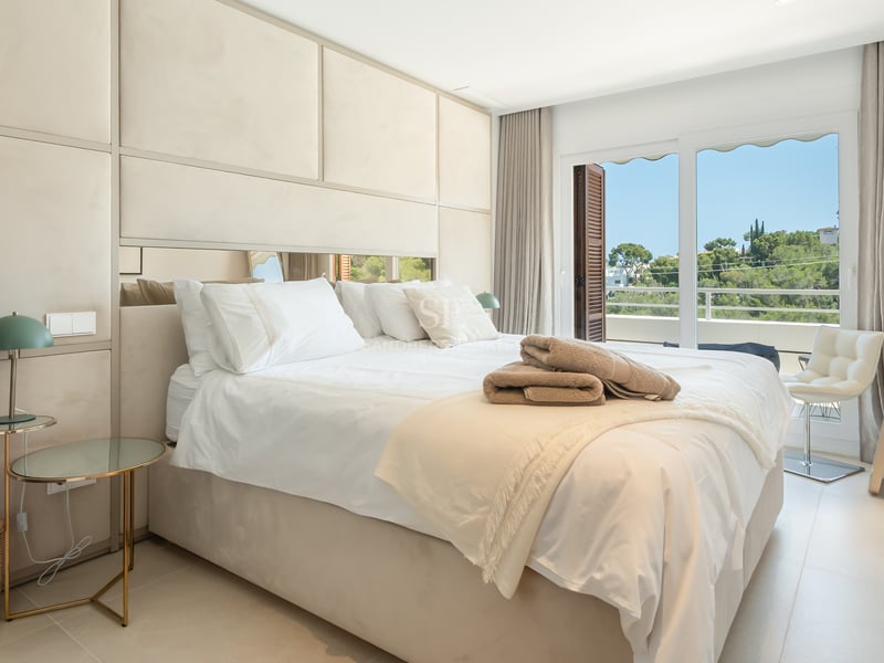 Chambre principale lumineuse avec tête de lit rembourrée, tables en verre et accès balcon vue jardin.