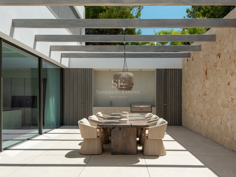 Terrasse moderne avec table en bois massif, chaises tressées, mur en pierre et poutres apparentes.