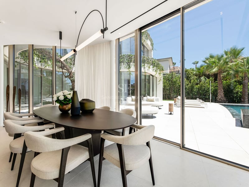 Salle à manger intérieure avec table en bois sombre et chaises beiges donnant sur une terrasse ensoleillée avec piscine.
