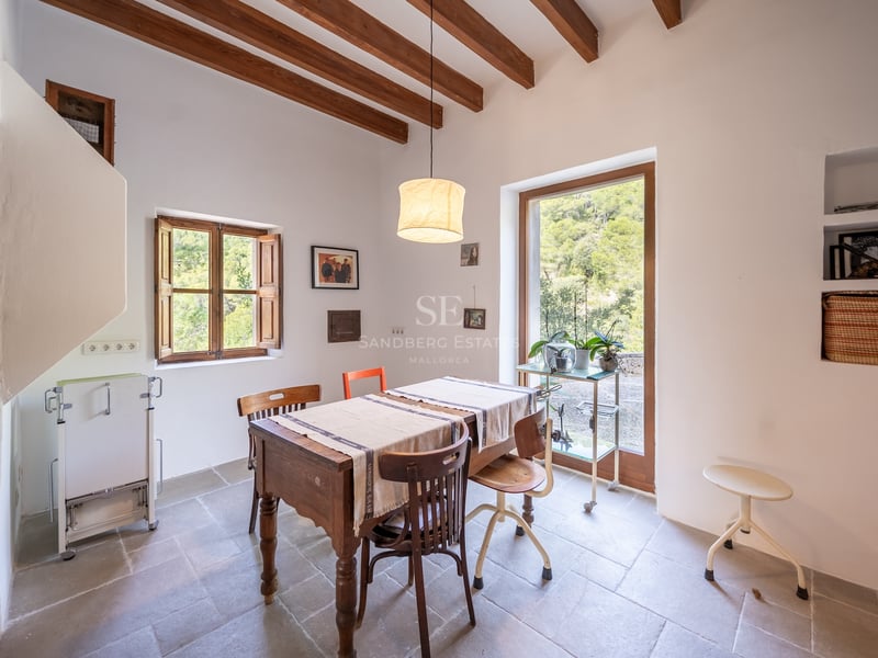 Salle à manger lumineuse avec table en bois, poutres apparentes et porte vitrée donnant sur la forêt.
