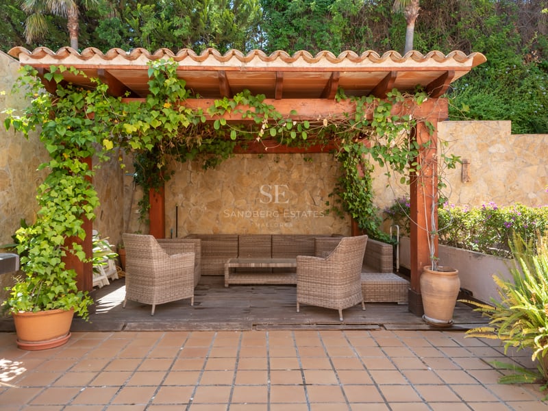 Terrasse extérieure avec pergola en bois, meubles en osier, murs en pierre et barbecue entouré de verdure.