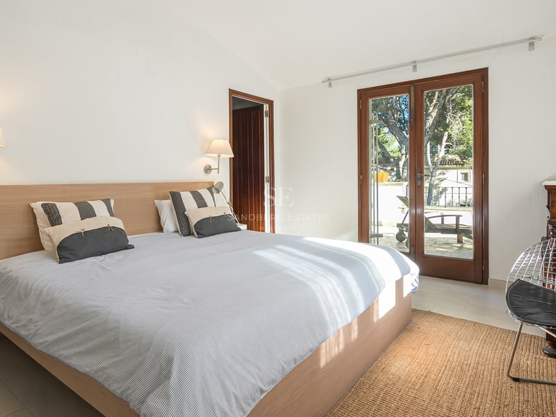 Chambre lumineuse avec lit en bois, murs blancs et portes vitrées ouvrant sur une terrasse ensoleillée.