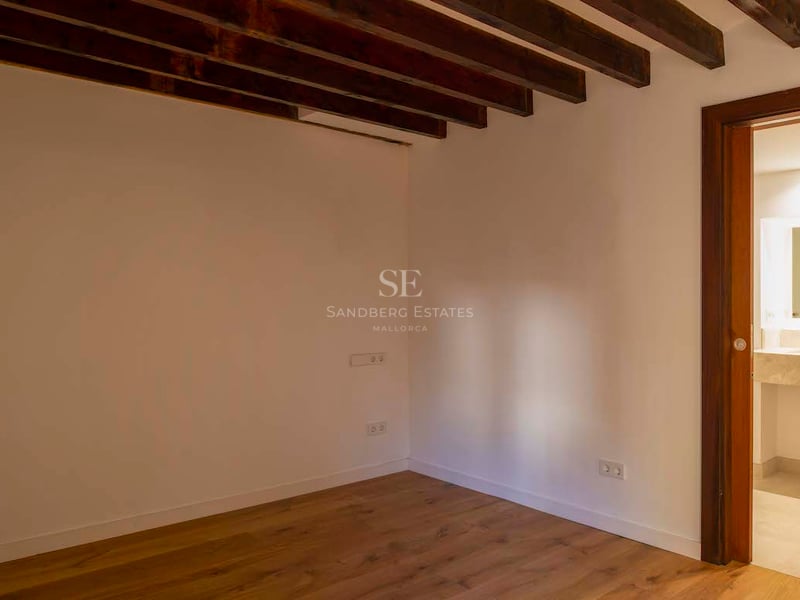 Chambre vide avec poutres de plafond en bois foncé, parquet et porte ouvrant sur une salle de bain moderne.