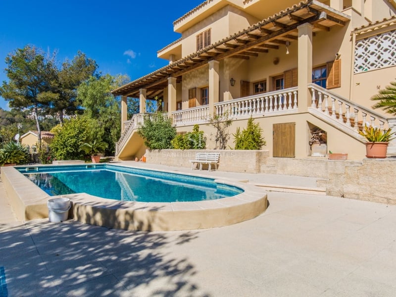 Grande piscine privée devant une villa méditerranéenne ocre avec terrasse et collines verdoyantes.