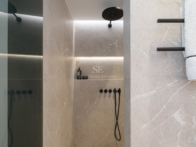 Douche avec carrelage en pierre beige, pommeau de pluie noir mat et niche éclairée sur un mur en pierre veinée.