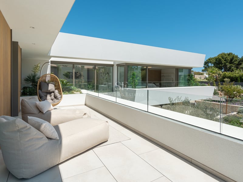 Vaste terrasse blanche avec chaises longues beiges, garde-corps en verre et vue sur un jardin paysager sous un ciel bleu.