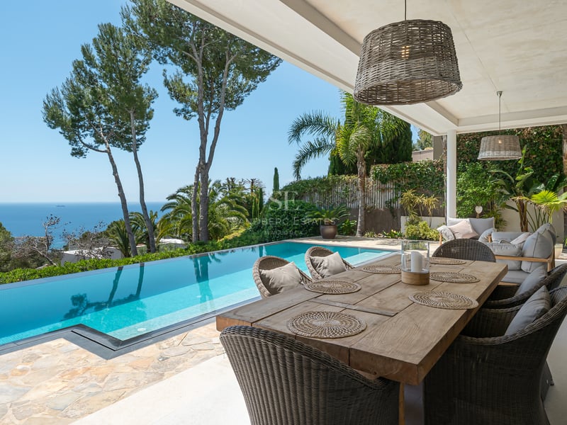 Terrasse avec table en bois à côté d'une piscine à débordement dominant la mer bleue et des pins.