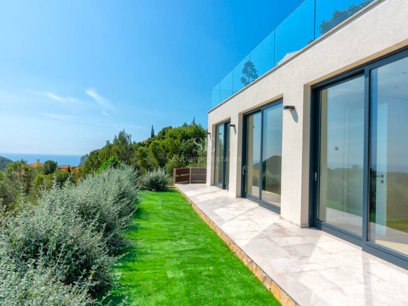 Villa blanche moderne avec grandes portes vitrées coulissantes, patio en pierre, pelouse et vue sur la mer et les montagnes.