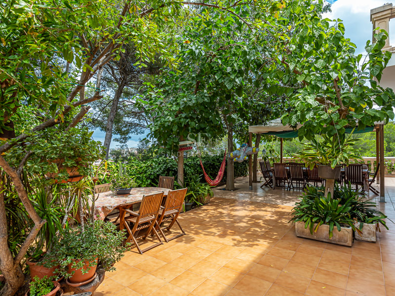 Grande terrasse carrelée entourée d'arbres matures avec deux espaces repas en bois et un hamac rouge à l'ombre.