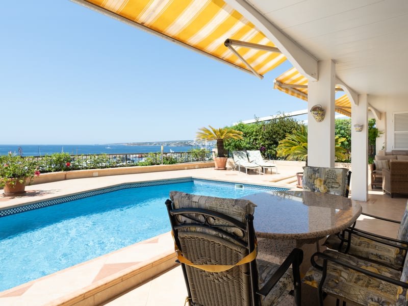 Terrasse avec piscine bleue, coin repas extérieur et stores jaunes offrant une vue imprenable sur la mer.