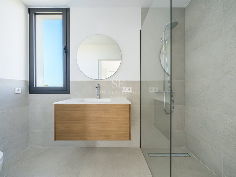 Salle de bain moderne avec carrelage gris, meuble en bois suspendu sous un miroir rond et douche à l'italienne vitrée.