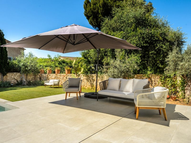 Terrasse ensoleillée avec canapé gris moderne, fauteuils et grand parasol à côté d'une piscine et d'un jardin.