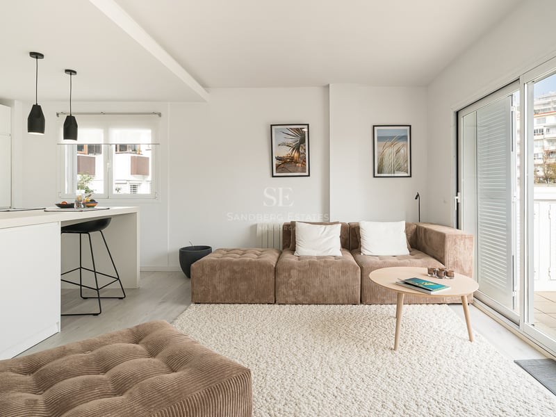 Salon lumineux avec canapé modulaire en velours côtelé marron, tapis blanc, îlot de cuisine et baies vitrées.