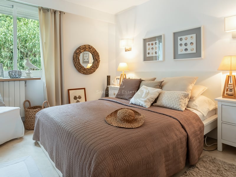 Chambre lumineuse avec lit matelassé brun, chevets blancs, miroir en osier et vue sur le jardin.