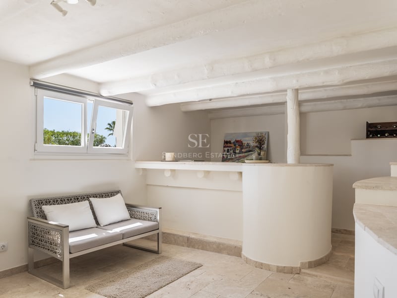 Intérieur blanc avec poutres en bois rustiques, sol en travertin, bar intégré et canapé moderne.