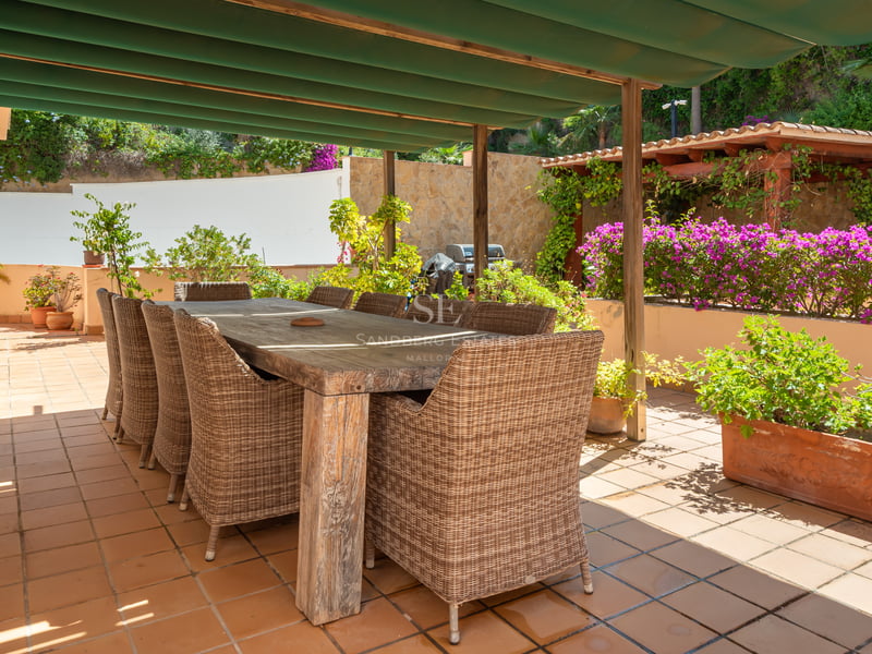 Grande table à manger en bois avec chaises en osier sur une terrasse en terre cuite ensoleillée sous un auvent vert.