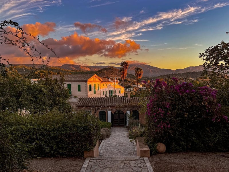 Vue panoramique d'une villa méditerranéenne blanche avec toit en terre cuite au coucher du soleil, entourée de jardins et de montagnes.