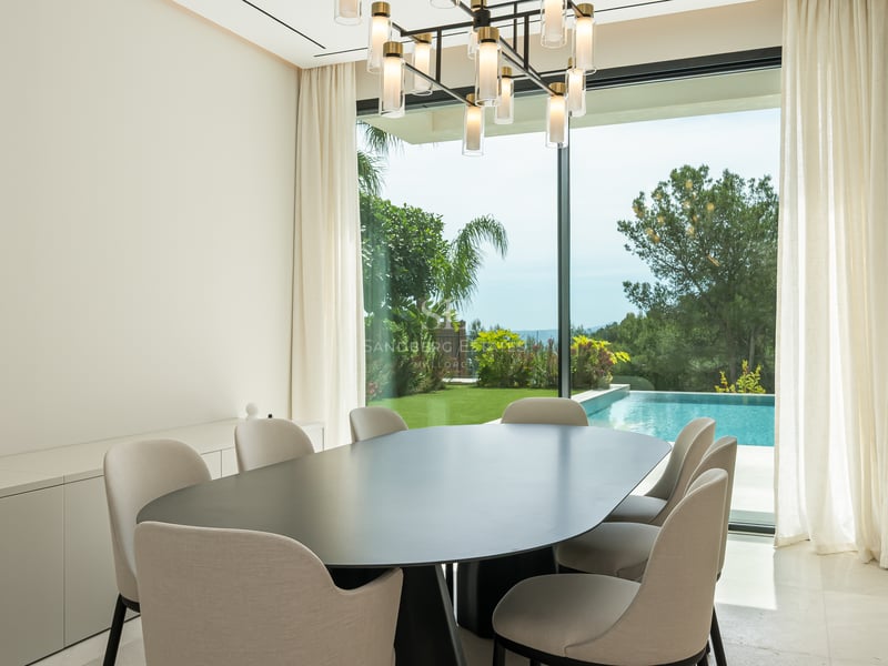 Espace dînatoire minimaliste avec table ovale noire et baies vitrées donnant sur un jardin avec piscine.