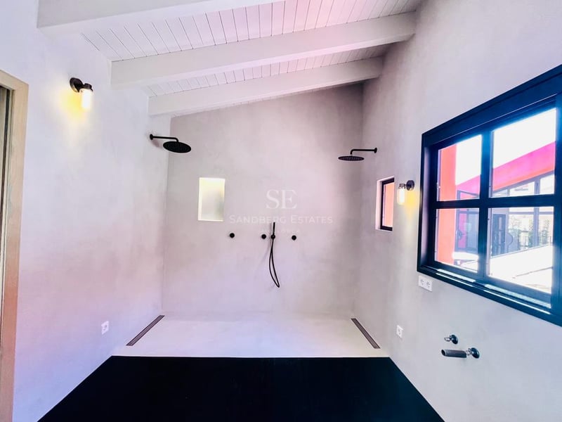 Douche minimaliste avec robinetterie noire mate, murs en microciment et poutres en bois blanc au plafond.