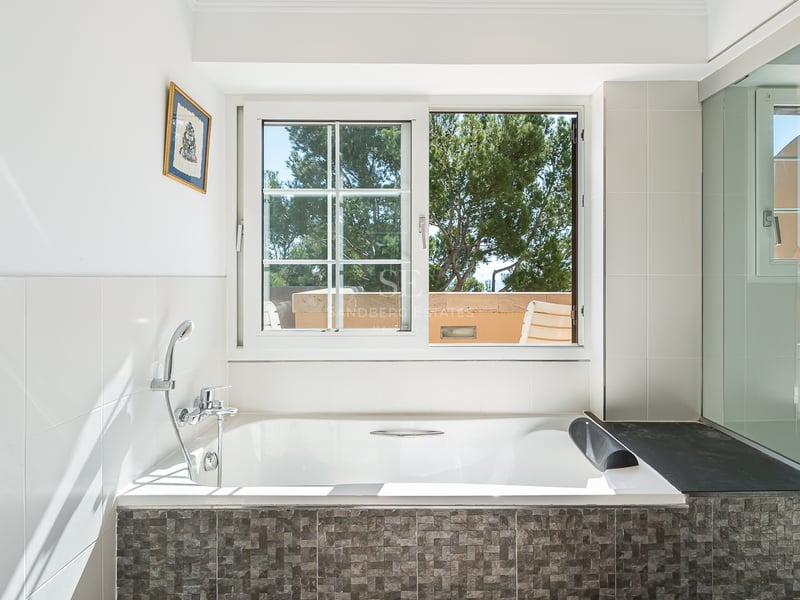 Salle de bain blanche avec baignoire encastrée, mosaïque grise et fenêtre donnant sur des pins méditerranéens.