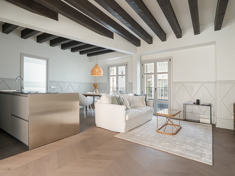 Salón moderno con vigas de madera oscura, suelo de parquet en espiga e isla de cocina de acero inoxidable.