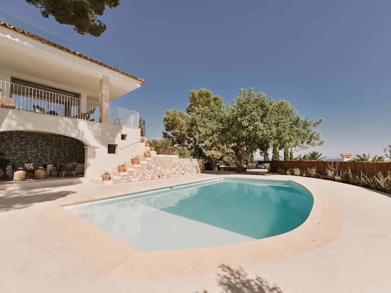 Piscine turquoise de forme arrondie bordée d'une villa blanche, d'escaliers en pierre et d'arbres verdoyants.