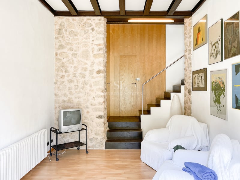Salon avec mur en pierre naturelle, poutres en bois sombre, escalier en ardoise et fauteuils blancs.