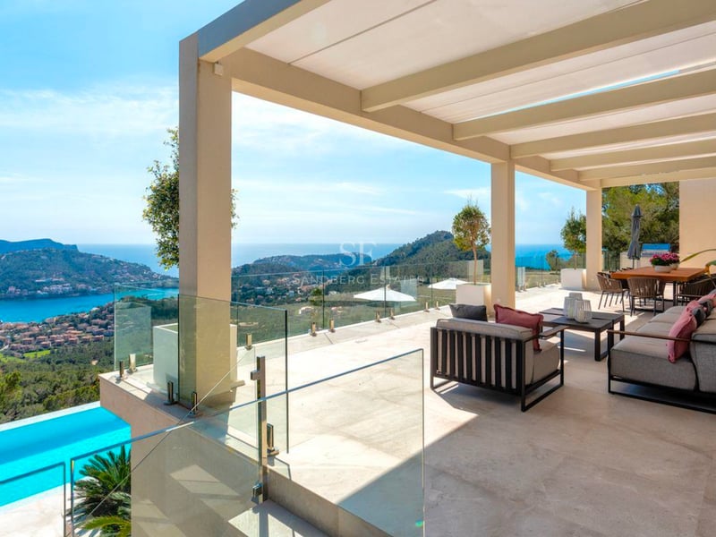 Grande terrasse moderne avec canapés gris, garde-corps en verre et vue imprenable sur une baie et des collines.