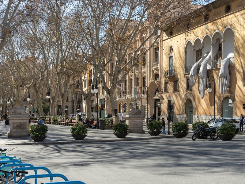 A tree-lined urban promenade with historic Mediterranean buildings, stone statues, and a sculpture of giant white hands.