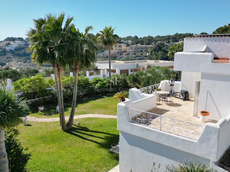 Vue d'en haut d'une villa blanche avec terrasse, jardin avec palmiers et vue sur la mer bleue et les falaises au loin.