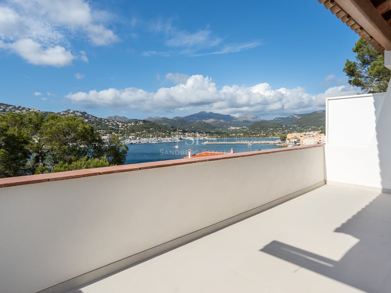 Grande terrasse blanche surplombant un port de plaisance avec voiliers, entourée de collines et montagnes.