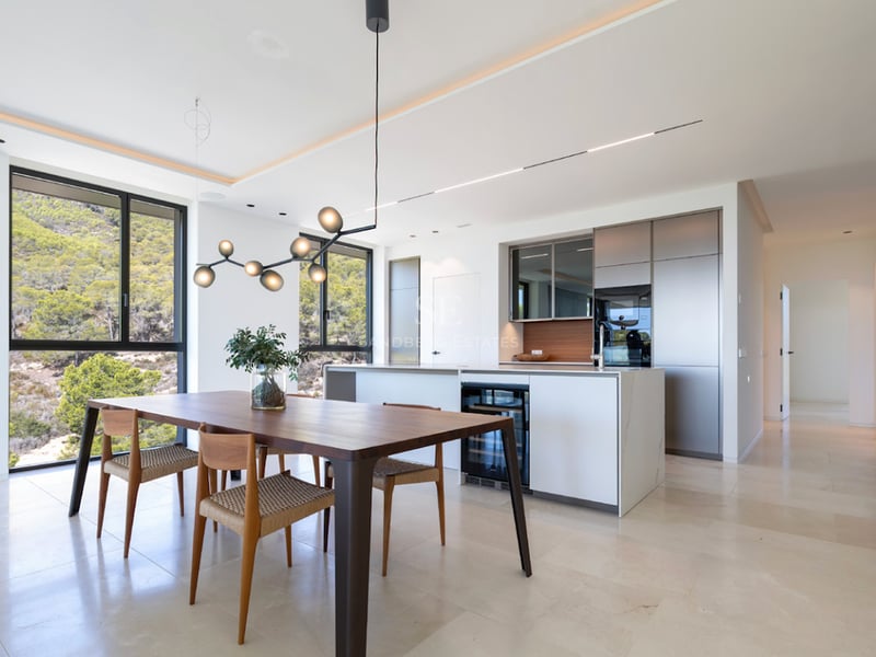 Cuisine moderne et salle à manger ouverte avec table en noyer, sol en pierre claire et vue sur les collines.