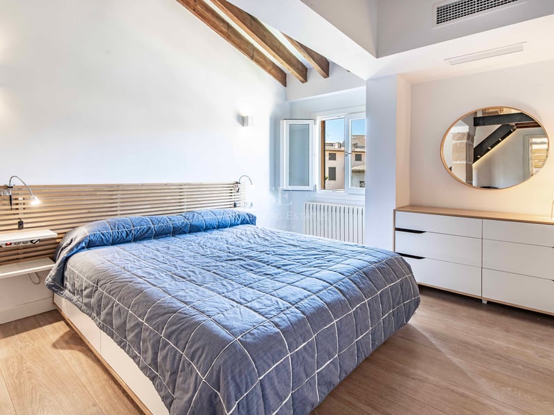 Chambre lumineuse avec murs blancs, poutres apparentes, couvre-lit bleu et un grand miroir rond sur la commode.