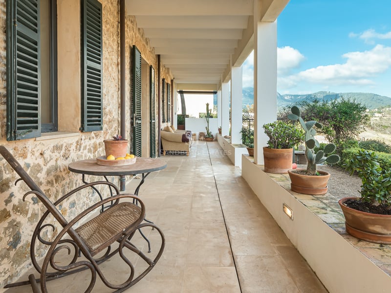 Longue terrasse couverte en pierre avec pots en terre cuite, volets verts et fauteuil à bascule face aux montagnes.